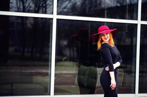 retrato de una chica pelirroja de moda con sombrero rojo y vestido negro con maquillaje brillante posado contra una ventana grande. filtros de instagram de estilo foto tonificado.