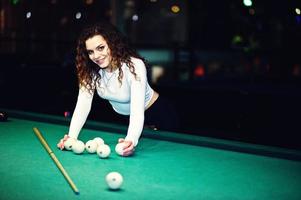 Young curly girl posed near billiard table. Sexy model at black mini mini skirt play russian snooker. Play game and fun concept. photo