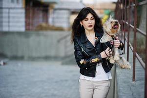 chica gitana morena con perro yorkshire terrier posó contra barandillas de acero. desgaste modelo en chaqueta de cuero y camiseta con adorno, pantalones. foto