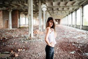 Portrait of young cute brunette girl wearing on black leather pants and white blouse posed on abandoned place. photo