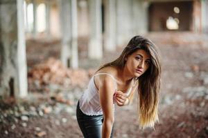 retrato de una joven morena linda con pantalones de cuero negro y blusa blanca posada en un lugar abandonado. foto