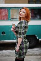 jovencita de pelo rojo con teléfono móvil y auriculares, vestida con un fondo de vestido a cuadros antiguo autobús minivan turquesa vintage. foto