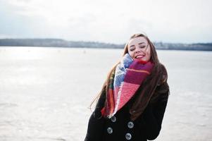 chica joven casual en abrigo negro, bufanda y sombrero contra el río congelado en el clima soleado de invierno. foto