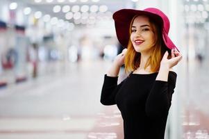 Fashion red haired girl wear on black dress and red hat posed at trade shopping center. Photo toned style Instagram filters.