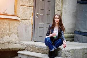 retrato de una joven elegante vestida con una chaqueta de cuero y jeans rotos con una taza de café. estilo de modelo de moda callejera. foto