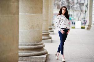 Young stylish brunette girl on shirt, pants and high heels shoes, posed near stone columns. Street fashion model concept. photo