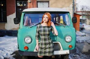 Young red haired girl posed on checkered dress background old retro cyan minivan. photo