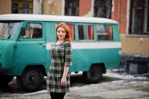 Young red haired girl posed on checkered dress background old retro cyan minivan. photo