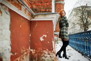 An outdoor portrait of a young pretty girl with red hair wearing checkered dress with girly backpacks standing on the brick wall background in winter day.