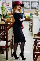 Portrait of fashion red haired girl on red hat and black dress with bright make up posed near table and chair salon. photo