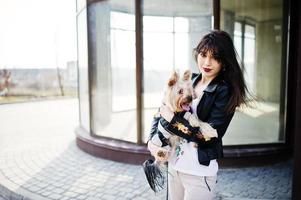 Brunette gypsy girl with yorkshire terrier dog posed against large windows house. Model wear on leather jacket and t-shirt with ornament, pants. photo