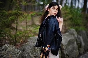 Brunette gypsy girl posed against stones on park. Model wear on leather jacket and t-shirt with ornament, pants. photo