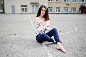 Young stylish teenage brunette girl on shirt, pants and high heels shoes, sitting on pavement and posed background school backyard. Street fashion model concept. photo