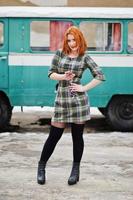 jovencita de pelo rojo con teléfono móvil y auriculares, vestida con un fondo de vestido a cuadros antiguo autobús minivan turquesa vintage. foto