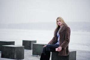 Stylish rich young blonde girl on fur coat with pink smartphone on hand background stone cubes against frozen lake on winter day. photo