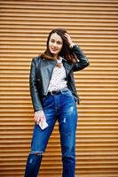 Portrait of stylish young girl wear on leather jacket and ripped jeans with mobile phone at hand background shutter texture. Street fashion model style.