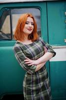 Young red haired girl posed on checkered dress background old retro cyan minivan.