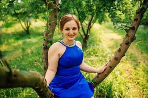 joven con sobrepeso en vestido azul posó jardín de primavera de fondo sentado en el árbol.