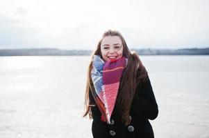 Casual young girl at black coat, scarf and hat against frozen river on sunny winter weather. photo