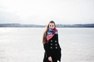 Casual young girl at black coat, scarf and hat against frozen river on sunny winter weather. photo