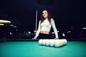 Young curly girl posed near billiard table. Sexy model at black mini mini skirt play russian snooker. Play game and fun concept. photo