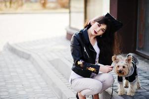 Brunette gypsy girl with yorkshire terrier dog posed against large windows house. Model wear on leather jacket and t-shirt with ornament, pants and shoes with high heels. photo