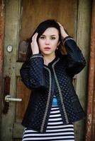 Brunette model girl at leather jacket posed background old wooden door of house. photo