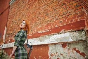 An outdoor portrait of a young pretty girl with red hair wearing checkered dress with girly backpacks standing on the brick wall background. photo