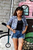 Portrait of beautiful teenage girl weared on checkered shirt and jeans shorts, against a wall with some element of graffiti. photo