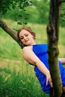 Young overweight girl at blue dress posed background spring garden sitting on tree. photo