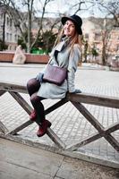 Young model girl in gray coat and black hat with leather handbag on shoulders posed against wooden beams at street of city. photo