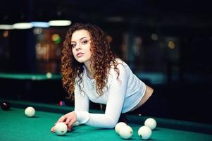 Young curly girl posed near billiard table. Sexy model at black mini mini skirt play russian snooker. Play game and fun concept. photo