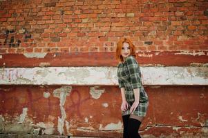An outdoor portrait of a young pretty girl with red hair wearing checkered dress standing on the brick wall background in winter day. photo