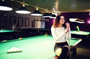 Young curly girl posed near billiard table. Sexy model at black mini mini skirt play russian snooker. Play game and fun concept. photo