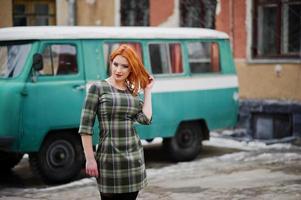 Young red haired girl posed on checkered dress background old retro cyan minivan. photo