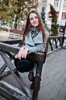 Young model girl in gray coat and black hat with leather handbag on shoulders posed against wooden beams at street of city. photo