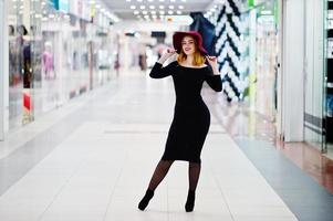 Fashion red haired girl wear on black dress and red hat posed at trade shopping center. Photo toned style Instagram filters.