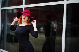 Portrait of fashion red haired girl on red hat and black dress with bright make up posed against large window. Photo toned style Instagram filters.