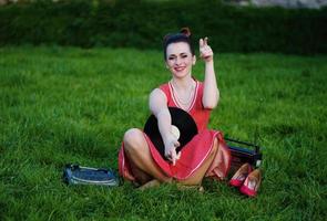 Portrait of young pinup girl wearing at retro vintage old-fashioned dress in peas sitting on grass wit retro radio and vinyl audio record in hands.