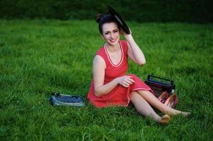 Portrait of young pinup girl wearing at retro vintage old-fashioned dress in peas sitting on grass wit retro radio and vinyl audio record in hands.