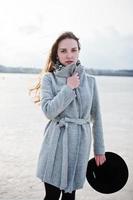 Young model girl in gray coat and black hat  posed against freeze lake. photo
