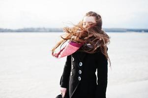 Casual young girl at black coat, scarf and hat against frozen river on sunny winter weather. photo