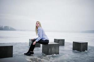 Stylish rich young blonde girl on blouse and leather pants with pink smartphone on hand background stone cubes against frozen lake on winter day. photo