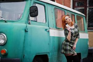 Young red haired girl lady with mobile cell phone and headphones, wearing on checked dress background old vintage turqoise minivan bus. photo