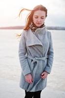 Young model girl in gray coat with red hair against freeze lake. photo