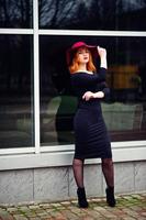 Portrait of fashion red haired girl on red hat and black dress with bright make up posed against large window. Photo toned style Instagram filters.