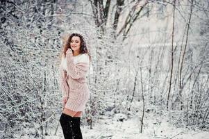 Curly brunette girl background falling snow, wear on warm knitted sweater, black mini skirt and wool stockings. Model on winter. Fashion portrait at snowy weather. Instagram toned photo. photo