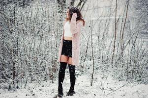 Curly brunette girl background falling snow, wear on warm knitted sweater, black mini skirt and wool stockings. Model on winter. Fashion portrait at snowy weather. Instagram toned photo. photo