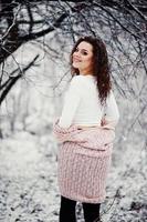 Back of curly brunette girl background falling snow, wear on warm knitted sweater, black mini skirt and wool stockings. Model on winter. Fashion portrait at snowy weather. Instagram toned photo. photo