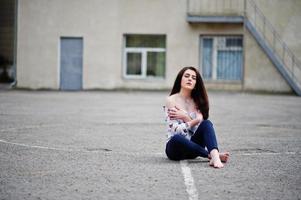 Young stylish teenage brunette girl on shirt, pants and high heels shoes, sitting on pavement and posed background school backyard. Street fashion model concept. photo